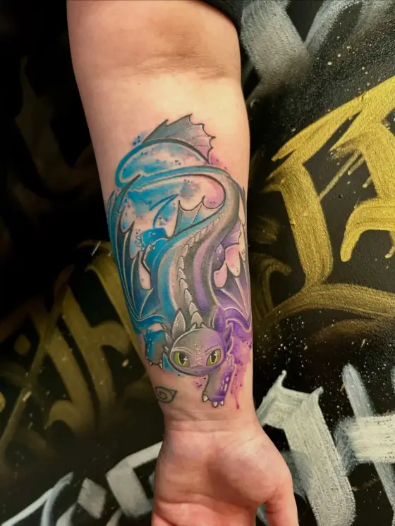 Tatouage couleur de Krokmou sur un avant-bras, devant le mur graffité noir et or du studio.